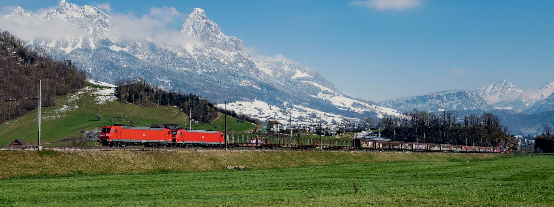 A freight train runs in front of a Swiss landscape with snow-covered peaks in the background