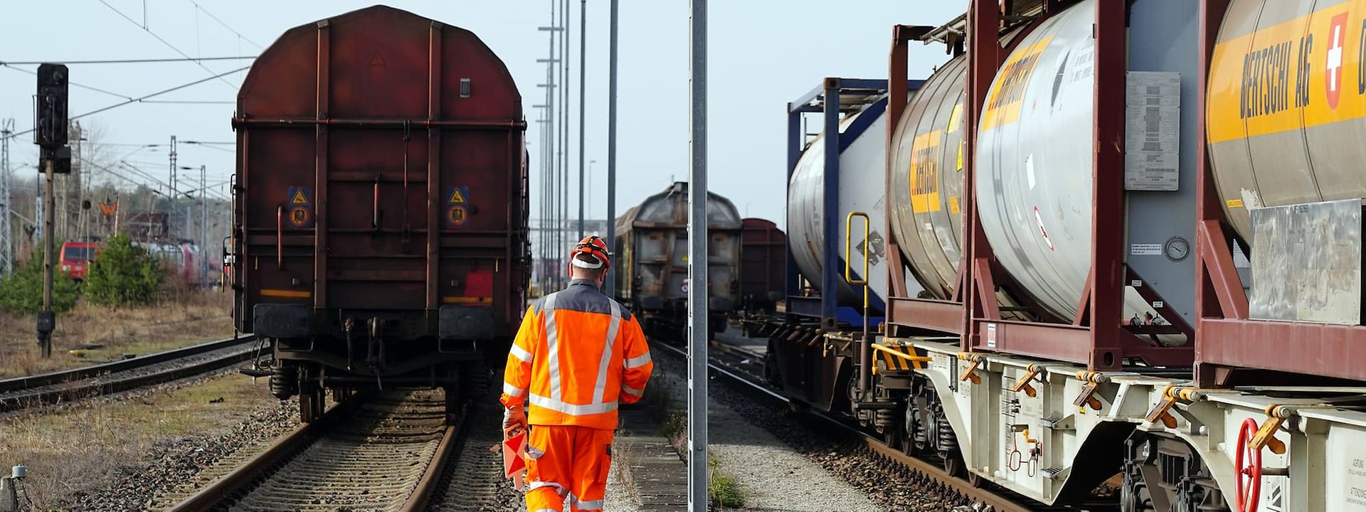 Rail worker in orange uniform walking between freight trains with tank containers.