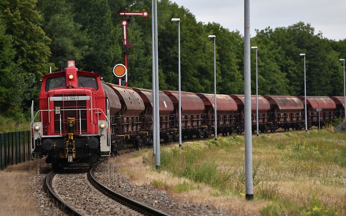 Red freight train with brown cargo wagons on a curved track surrounded by trees, passing signal lights.